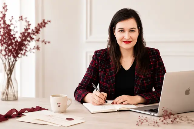 Grafička píše do skicáku a pracuje na návrhu svatebního oznámení; na stole má notebook, hrnek, obálky s pečetí a dekorativní větvičky.